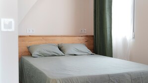 Un letto singolo con testiera in legno, due cuscini chiari e copripiumino chiaro in una stanza con soffitto spiovente e finestra con tenda verde.