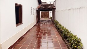 A narrow, wet alleyway with a tiled floor, flanked by white walls and a wooden gate at the end. - Beach House Unid. 2 Casas Praia Guarujá (Guarujá)