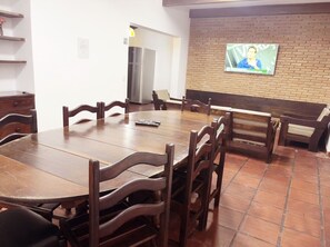 A dining area with a wooden table and chairs, a bench, a TV mounted on a brick wall, and a refrigerator in the background. - Beach House Unid. 2 Casas Praia Guarujá (Guarujá)