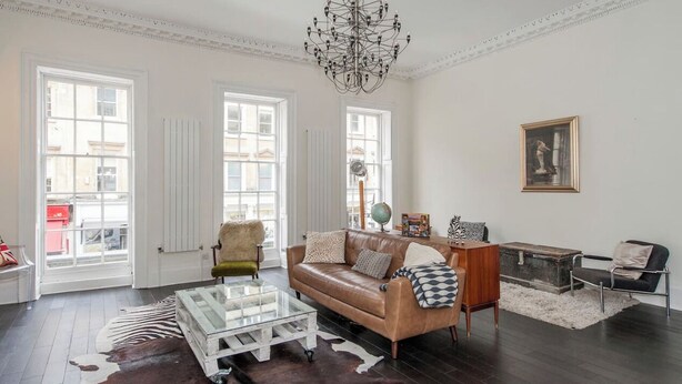 A spacious living room with a leather sofa, a glass coffee table, a chandelier, and a view through large windows. - Breathtaking Flat in Central Bath !! (Bath)