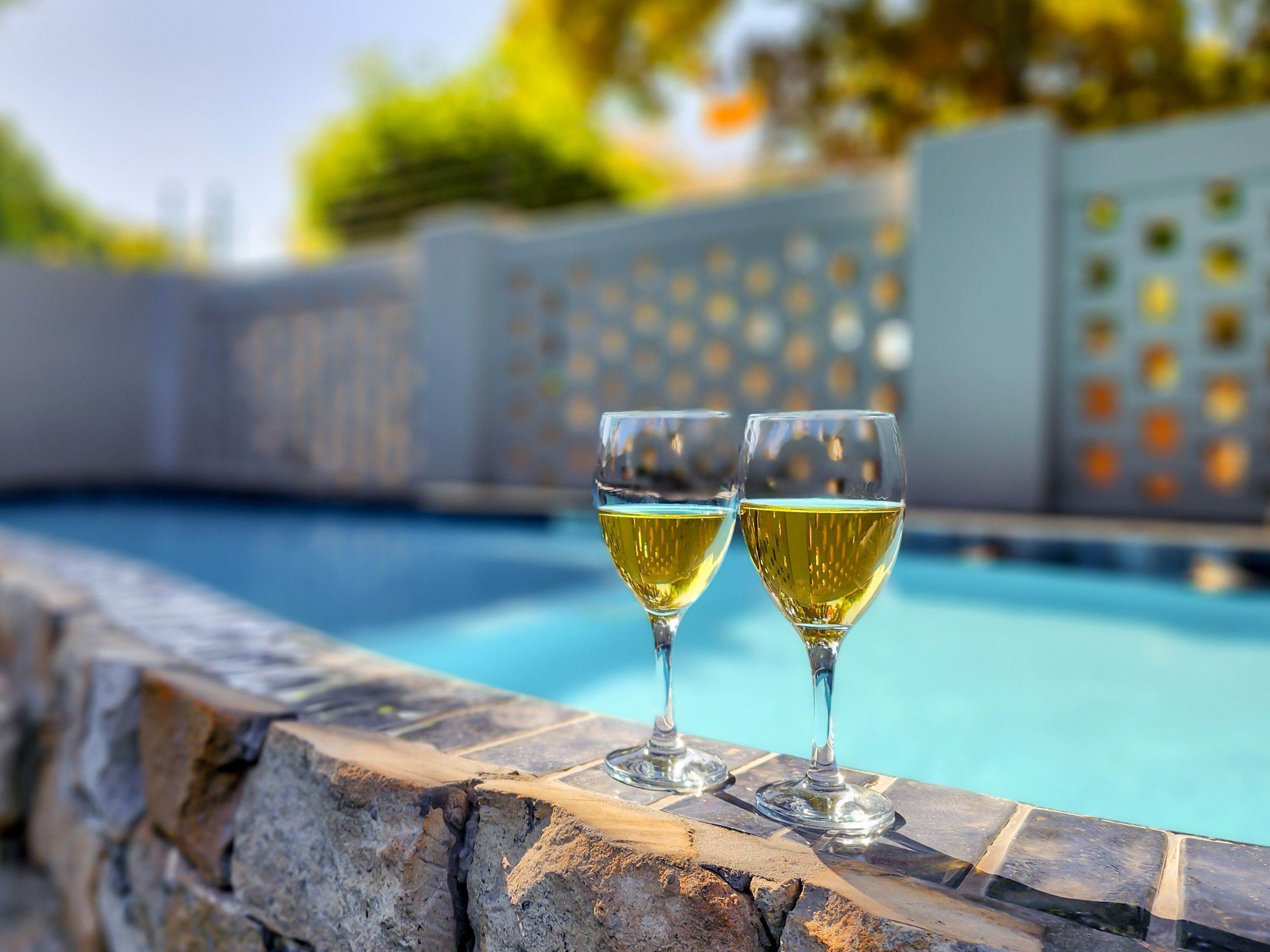 Two glasses of white wine on a stone ledge by a pool.