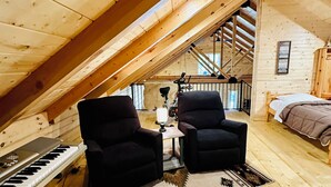A cozy attic room with wooden floors, two black armchairs, a wooden table, a keyboard, and a bed with a brown blanket. - Pine Cabin on Gilmore Lake (Minong)
