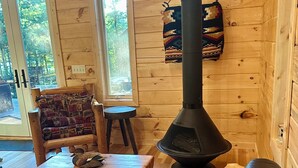 A cozy cabin interior with a black wood-burning stove, a leather sofa, a wooden chair, and a small table with a bird on it. - Pine Cabin on Gilmore Lake (Minong)