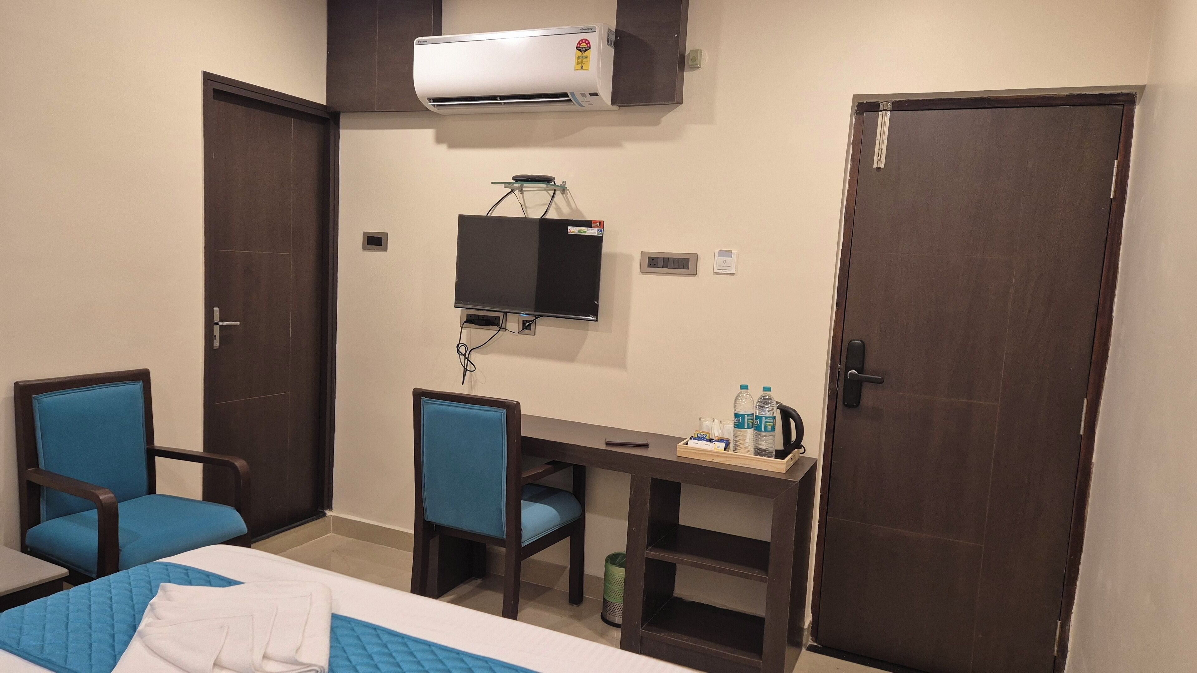 Una habitación de hotel con una cama, dos sillas azules, un escritorio con una computadora y botellas de agua, un aire acondicionado y una puerta cerrada.