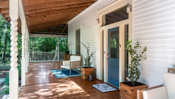 A covered porch with a wooden ceiling, white railings, and a blue door. There are two potted plants and a wicker chair on the porch. - Lavender Hill Bed & Breakfast (Athens)