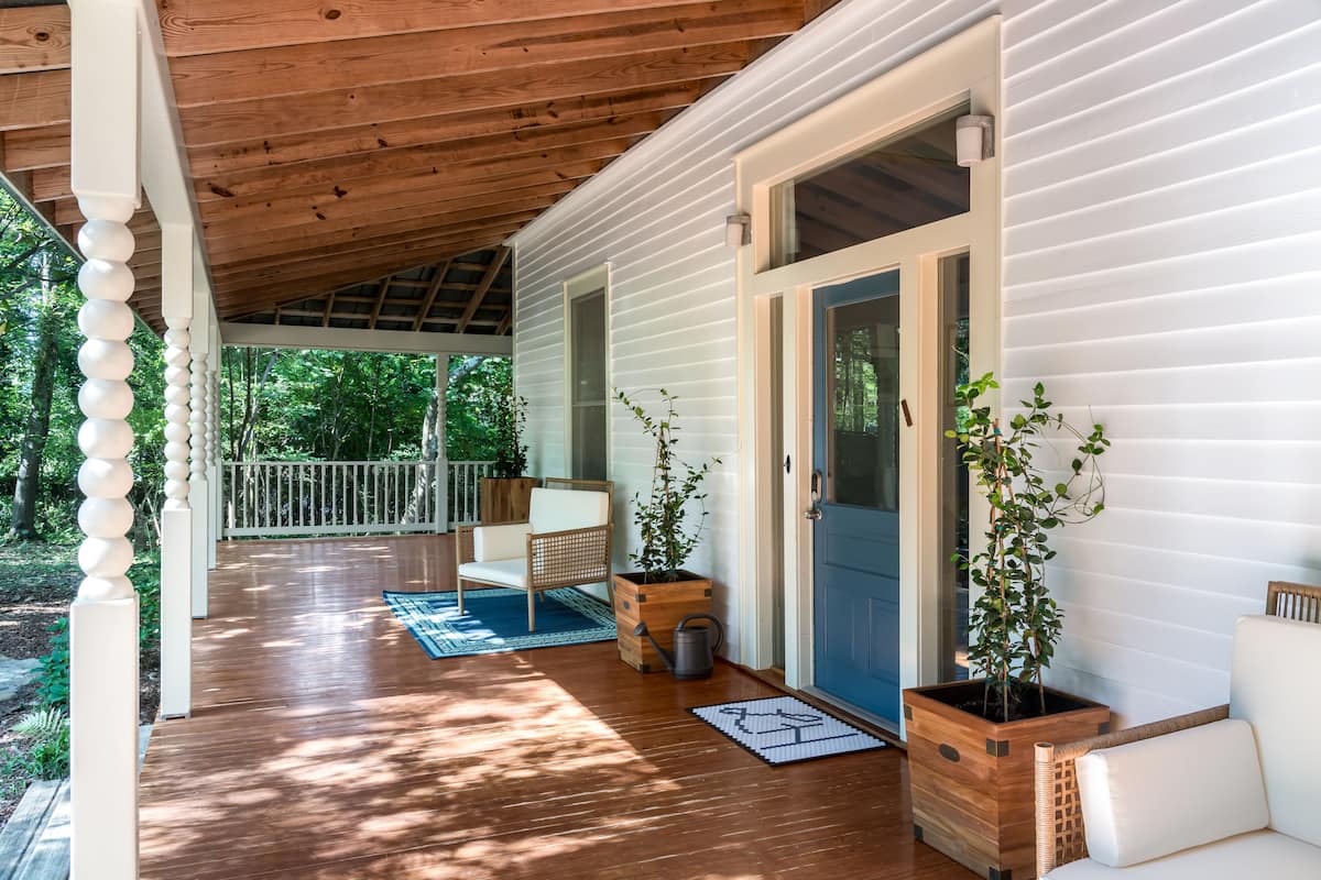 A covered porch with a wooden ceiling, white railings, and a blue door. There are two potted plants and a wicker chair on the porch.