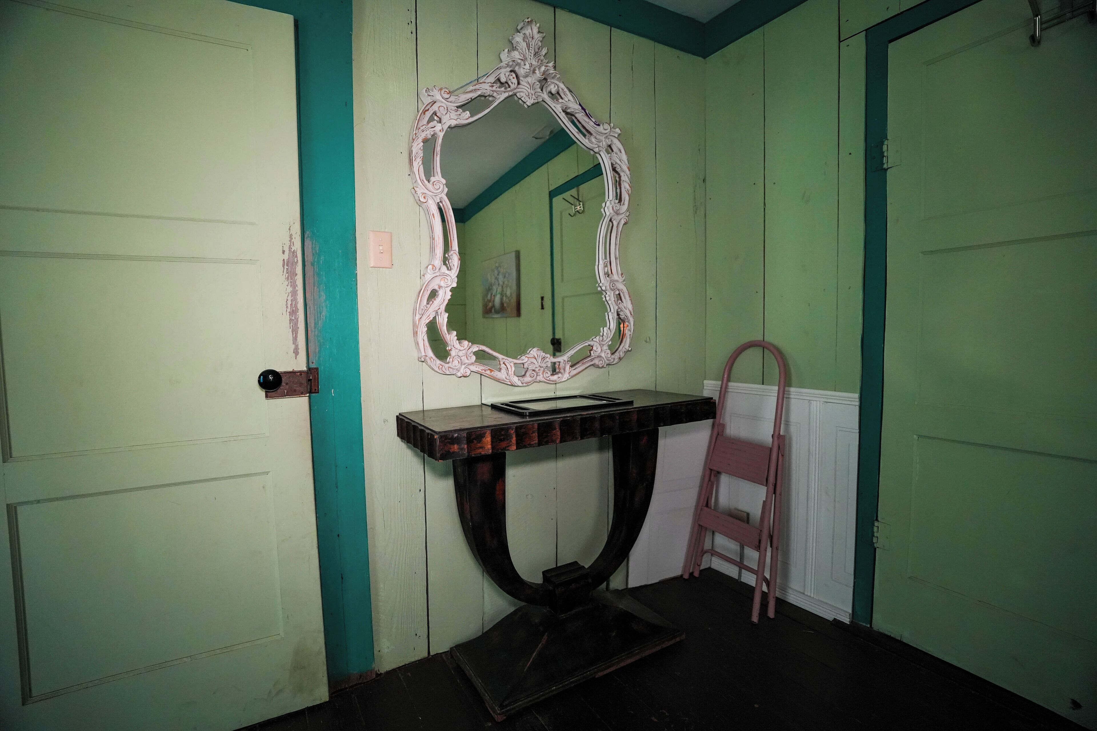 A room with a teal and white color scheme, featuring a large ornate mirror, a wooden console table, and a ladder against the wall.