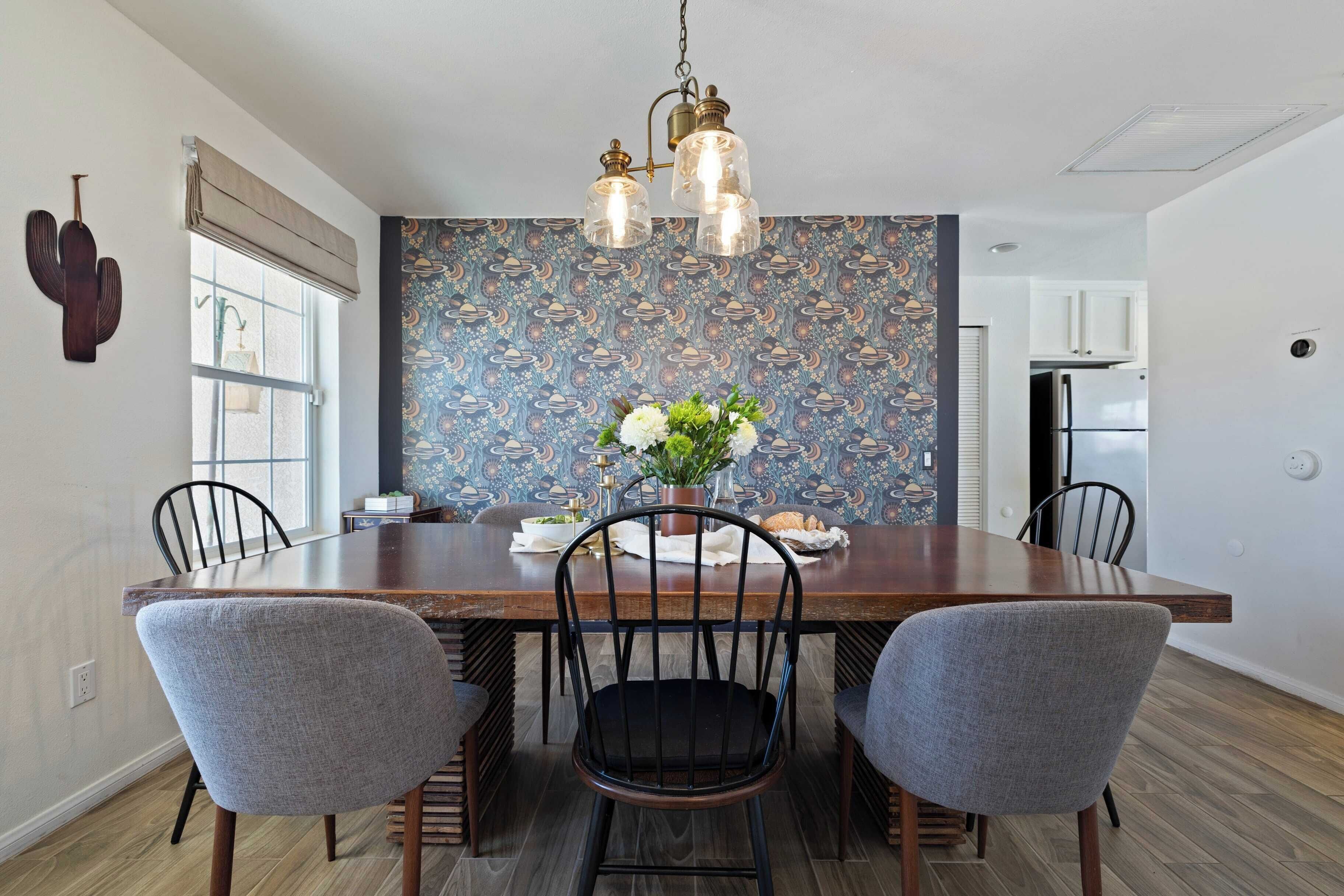 A dining area with a wooden table, four chairs, a chandelier, and a wall with a space-themed wallpaper.