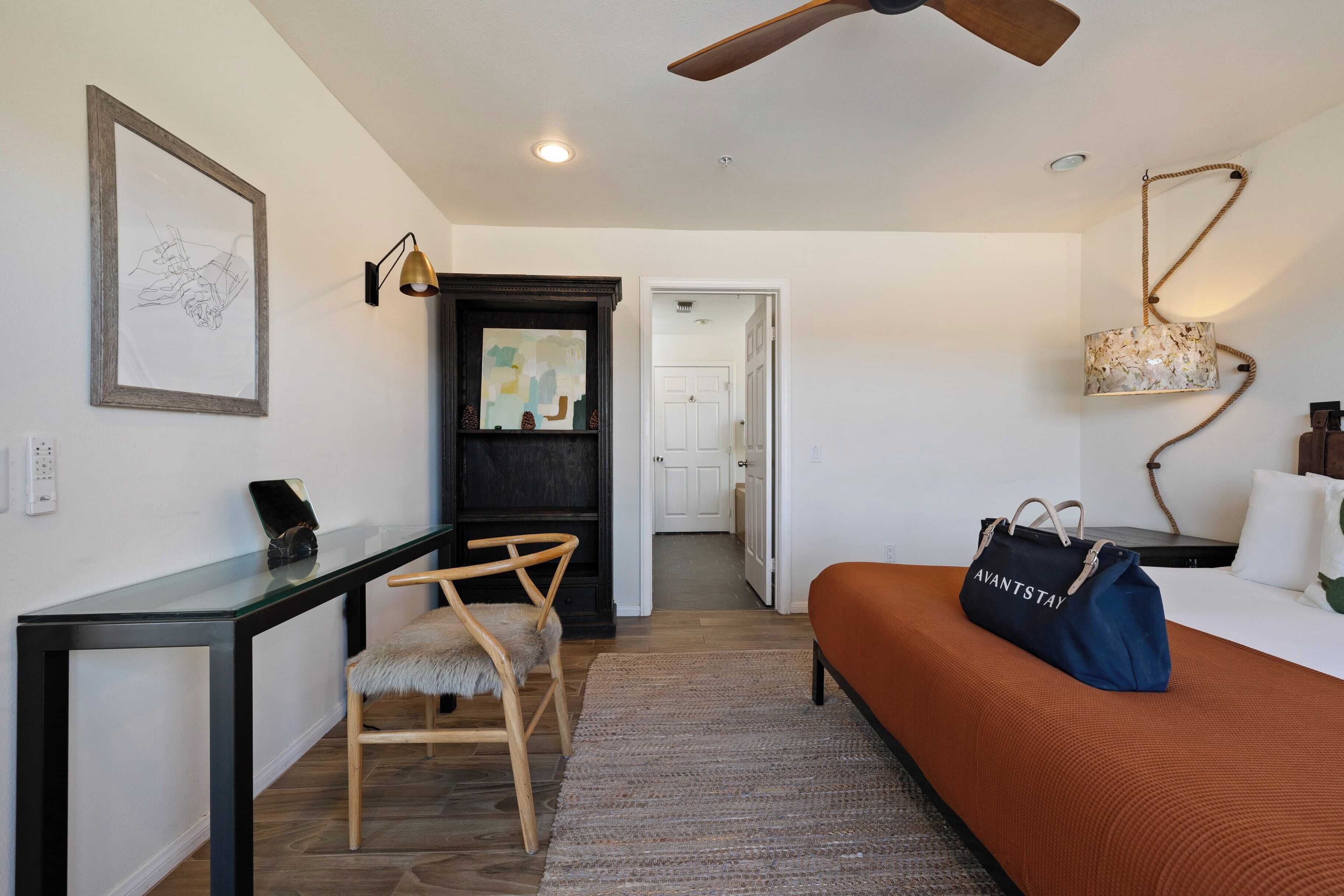 A hotel room with a bed, a desk, a chair, a hanging bag, and a door leading to another room.