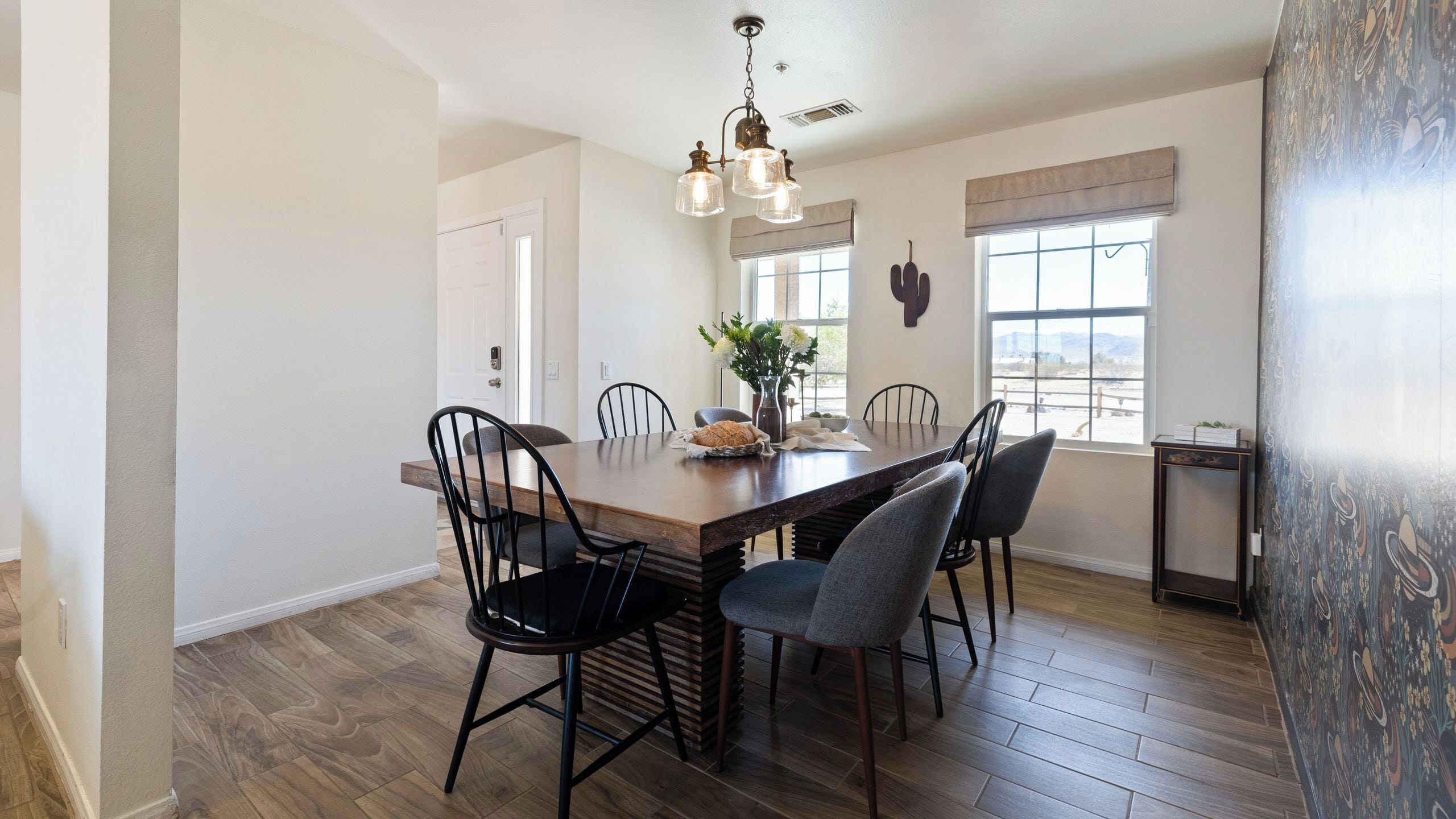 A dining area with a wooden table, four chairs, a vase with flowers, and a hanging light fixture.