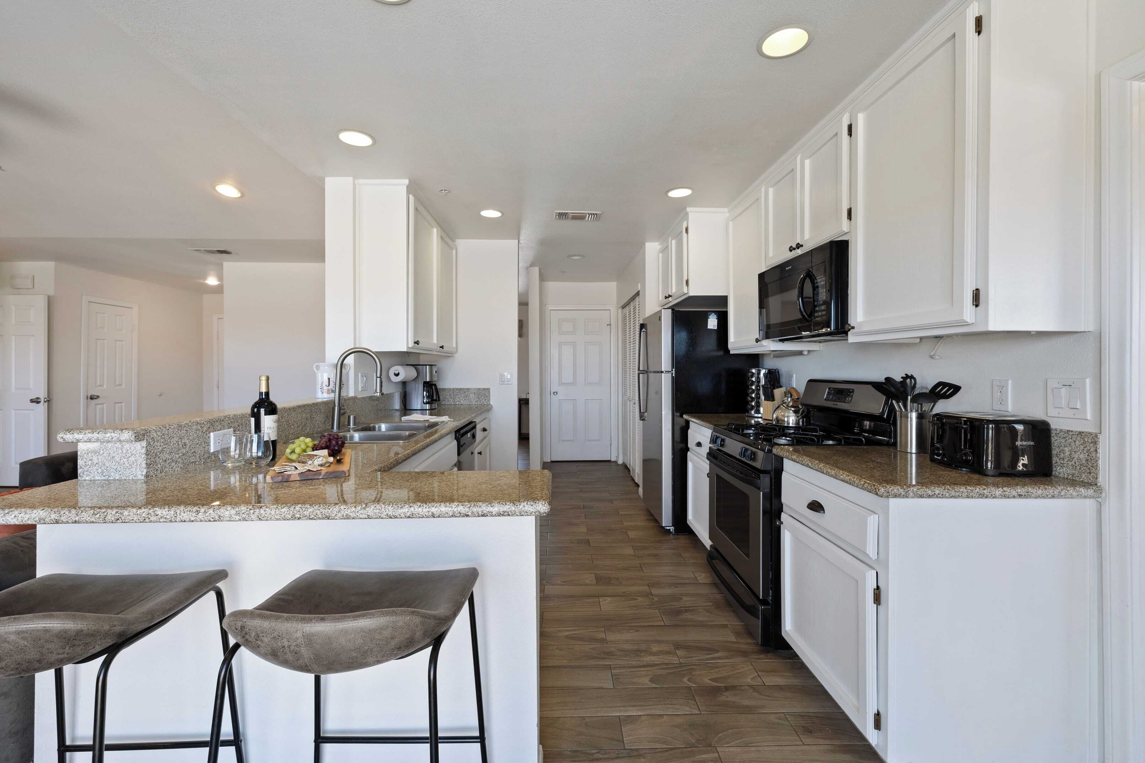 A modern kitchen with white cabinets, granite countertops, and stainless steel appliances. There are two bar stools at the island, and a refrigerator with a built-in microwave above the stove.
