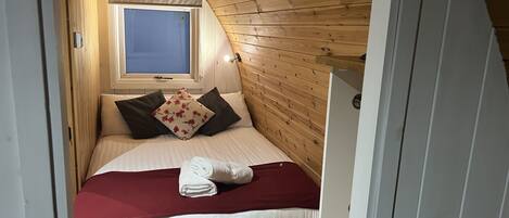 Une chambre avec un plafond en pente, des murs en bois, une fenêtre, un lit avec des oreillers et une serviette enroulée.