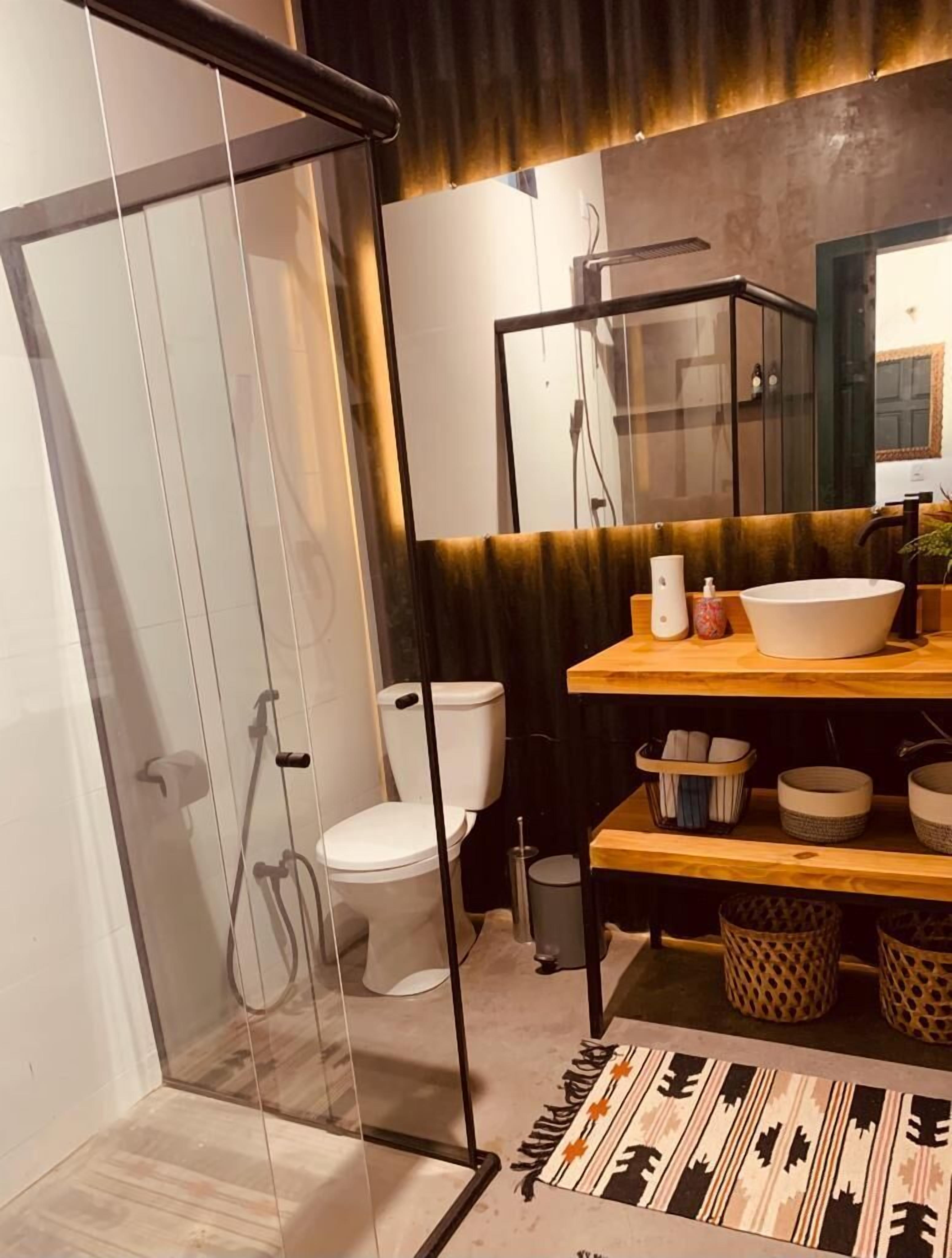 A modern bathroom with a glass shower enclosure, a white toilet, a wooden vanity with a large sink, and a mirror above the vanity.