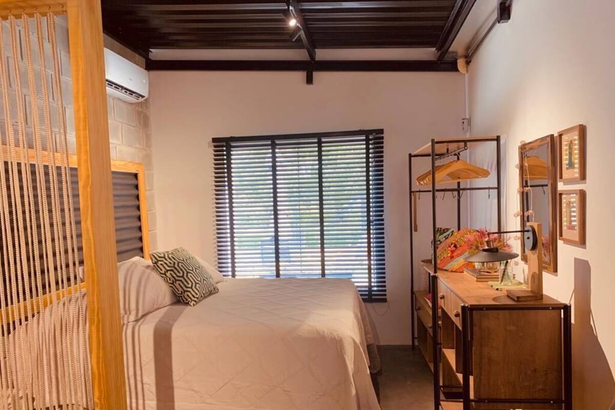 Een slaapkamer met een bed, een bureau en een stoel. Er is een raam met zonwering, een hanglamp en een wandplank met decoratieve objecten.
