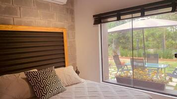 Quarto com uma grande janela, cama com almofadas estampadas e vista para um pátio externo.