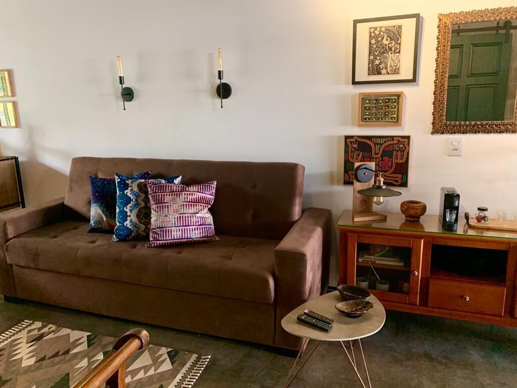 Un salón con un sofá marrón, cojines estampados, una mesita redonda con un control remoto y un aparador de madera con adornos.