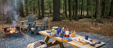 A picnic setup with a fire pit, wooden table, and chairs in a forest.