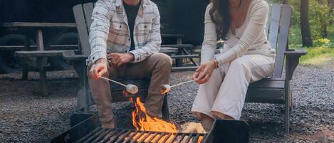 Two people sitting on chairs near a campfire in a forested area with a cabin in the background.