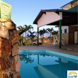 A swimming pool with a stone wall, a covered patio area, and palm trees in the background. - POUSADA SÃO JUDAS TADEU (Guaratingueta)