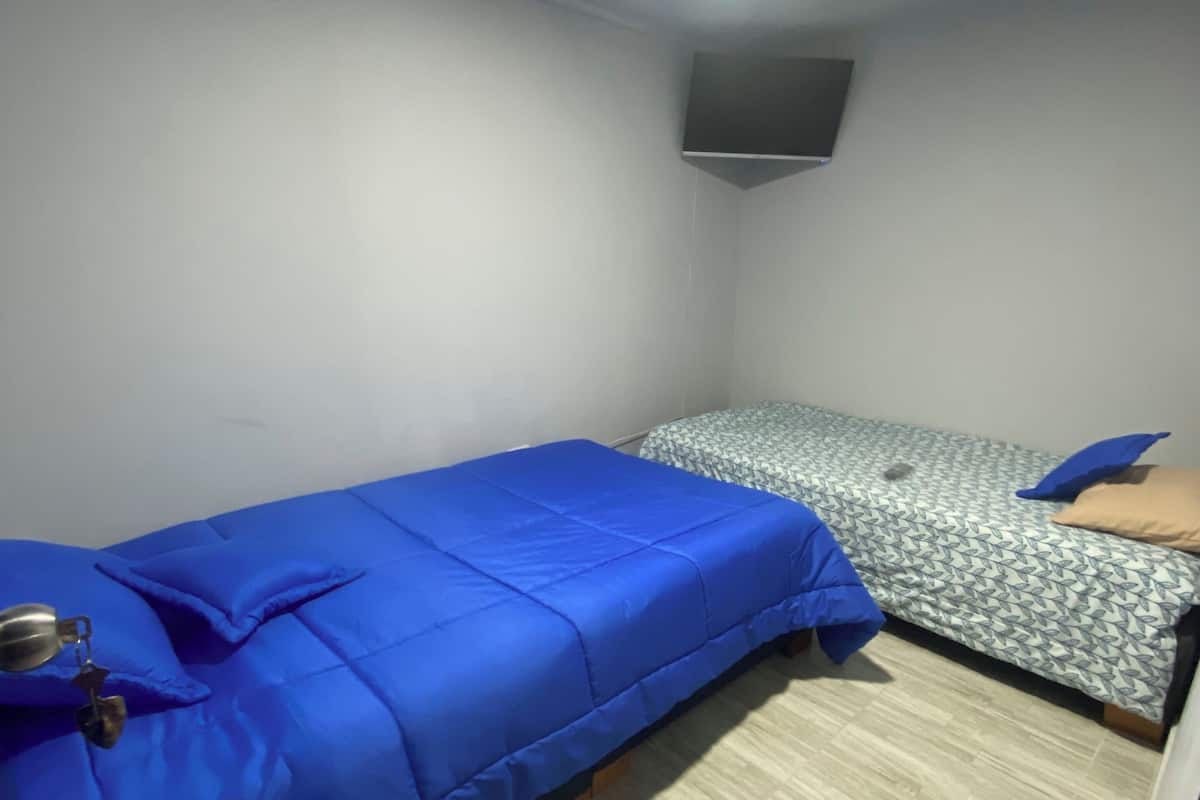 A small, single-room accommodation with two beds, one with a blue cover and the other with a patterned cover. A flat-screen TV is mounted on the wall above the beds.