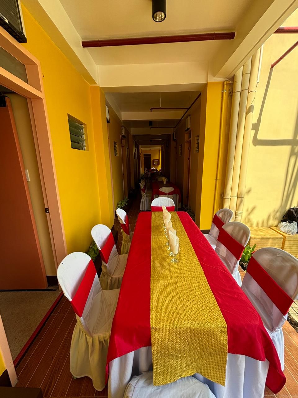 A long dining table with a red and gold table runner, white chairs with red sashes, and a yellow wall in the background.