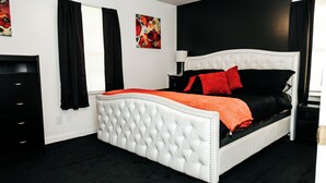 Ein Schlafzimmer mit einem weißen, gepolsterten Kopfteil, rot-schwarzer Bettwäsche, einer schwarzen Kommode und einem Fenster mit schwarzen Vorhängen.