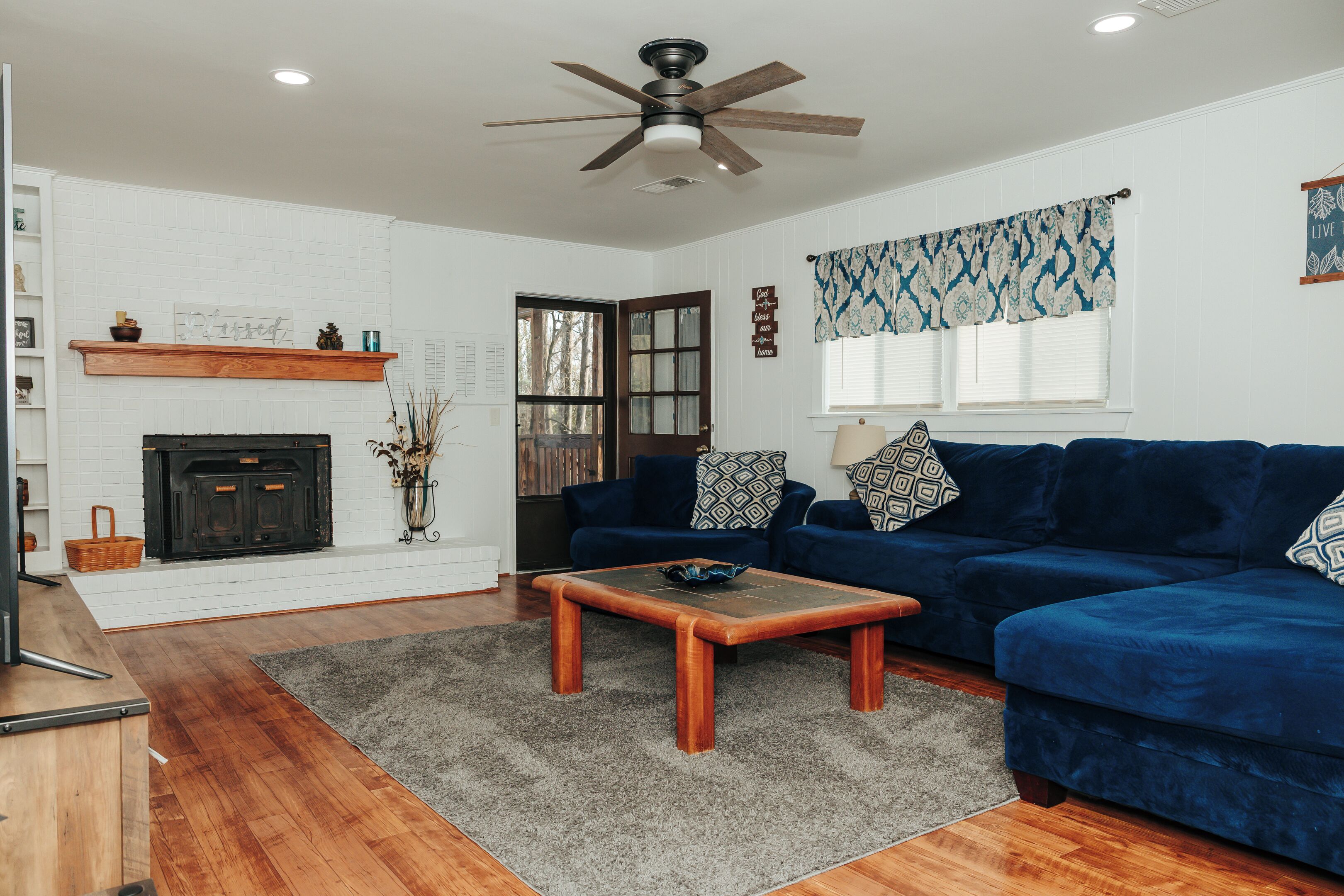 Una sala de estar con chimenea, sofá azul, mesa de centro de madera y ventilador de techo.