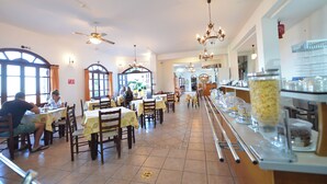 A restaurant with multiple tables and chairs, ceiling fans, and hanging chandeliers. - Ikaros Star Hotel (Ikaria)