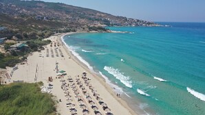 A beach with rows of sunbeds and umbrellas, a clear blue sea, and buildings on a hillside in the background. - Ikaros Star Hotel (Ikaria)
