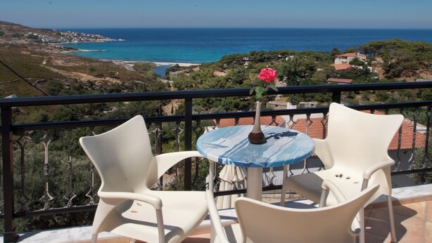 A balcony with two white chairs and a small round table, overlooking a coastal landscape with buildings and trees. - Ikaros Star Hotel (Ikaria)