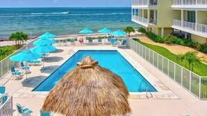 A pool area with a thatched roof, lounge chairs, and umbrellas, overlooking the ocean and a hotel building.