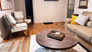 Uma sala de estar com piso de madeira, uma mesa de centro redonda com um cesto trançado, um sofá bege com almofadas amarelas e cinzas, uma cadeira de madeira e um quadro na parede.
