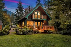 Une cabane en rondins à deux étages, dotée d’un balcon, entourée d’une végétation luxuriante et avec une chaîne de montagnes en toile de fond.