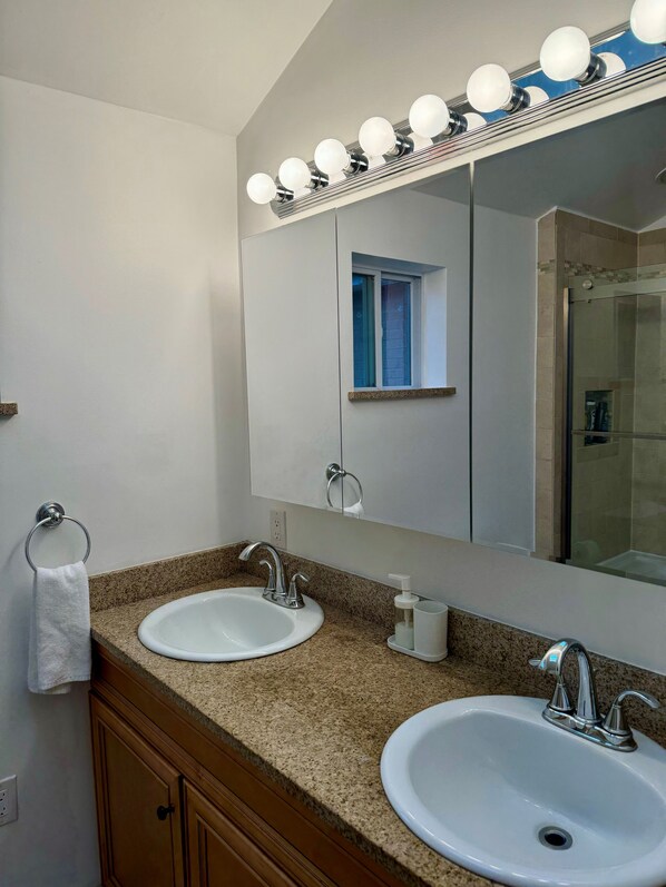 A bathroom with two sinks, a mirror with lights, a window, and a shower.