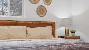 A bedroom with a wooden headboard, a bed with beige pillows, a bedside table with a lamp and a plant, and three woven wall decorations.