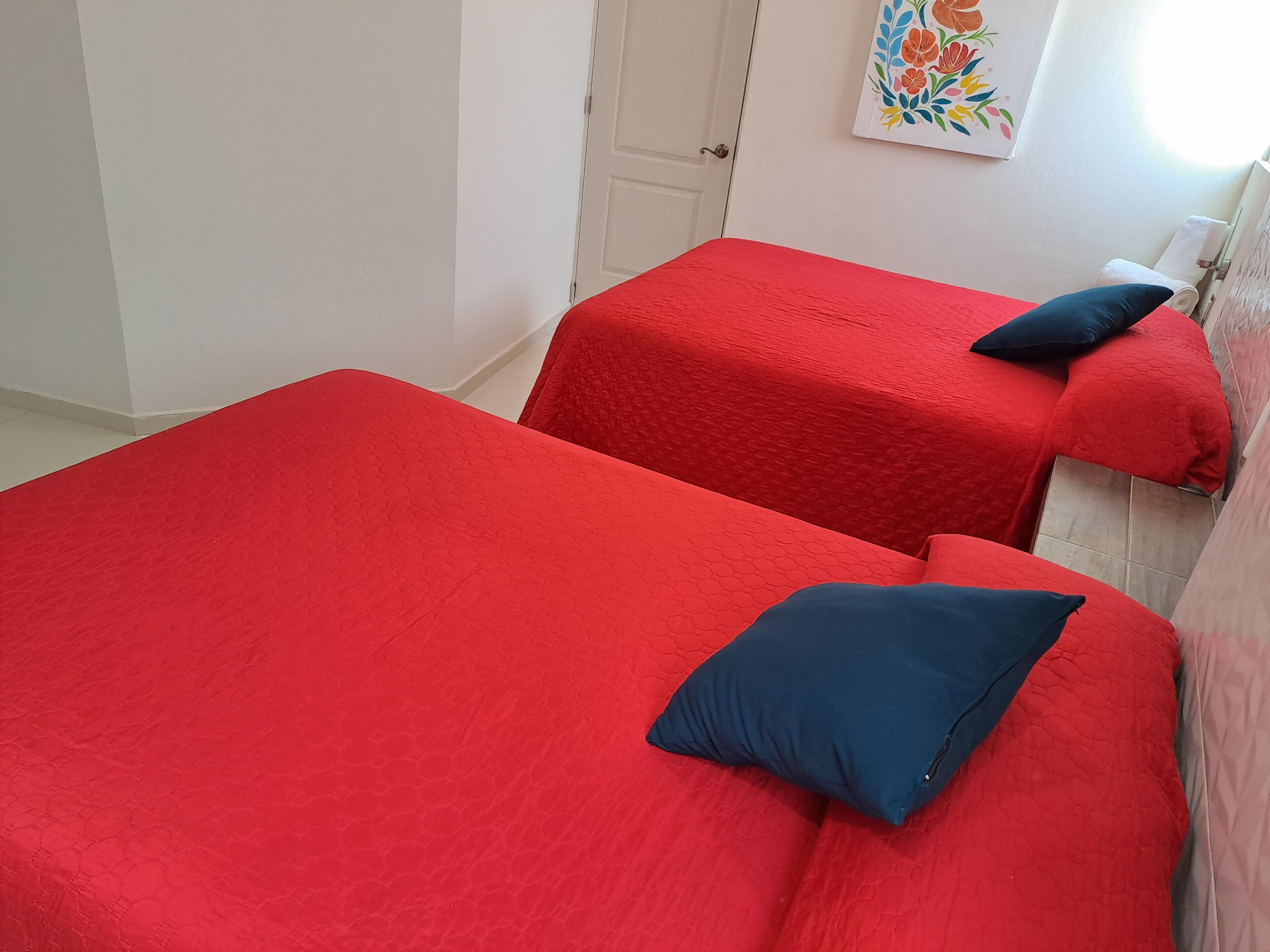 Two single beds with red bedding and dark blue pillows in a room with white walls and a door.