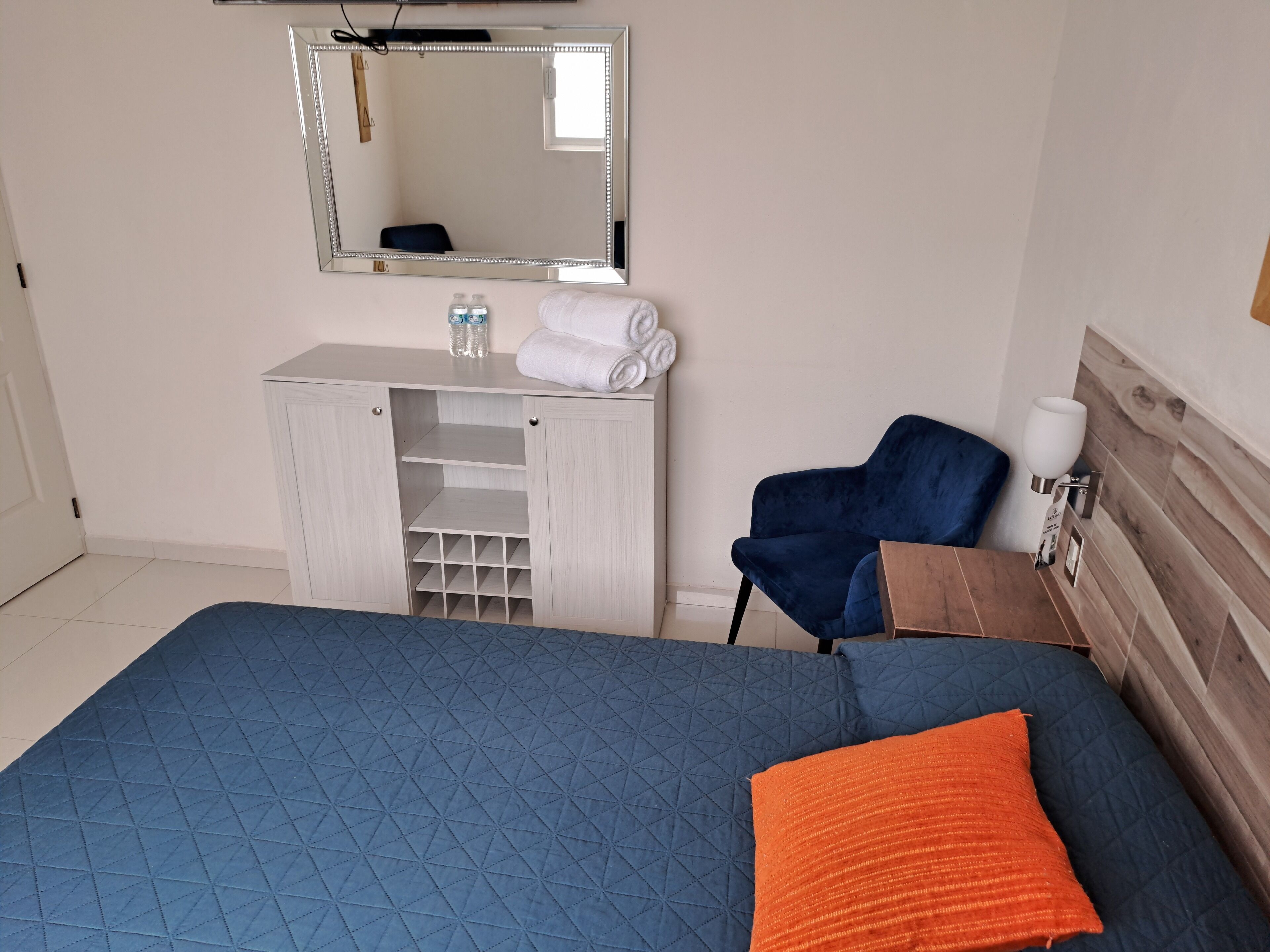A bedroom with a bed, a blue chair, a wooden cabinet, and a mirror.