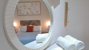 Un espejo circular refleja una cama con almohadillas blancas y naranjas, una colcha azul y un cabecero de madera. Una toalla blanca está doblada a un lado.