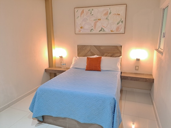 A neatly made bed with a blue quilt, two bedside tables with lamps, and a floral painting above the bed. - Hotel Anturio (Dolores Hidalgo)