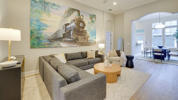 A spacious living room with a large painting of a train, a gray sectional sofa, a round coffee table, and a dining area with a blue table and white chairs. - Townhome w/ Patio < 5 Mi to UF Campus! (Gainesville)