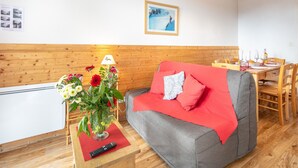 A cozy room with a grey sofa, red cushions, a wooden table with a vase of flowers, and a dining area with wooden chairs and a table set for two. - Hermine - apt 6p, vue montagne (Chamrousse)