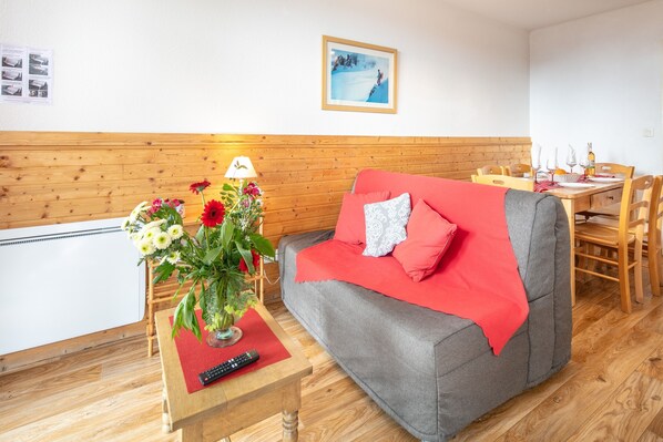 Ein gemütliches Zimmer mit grauem Sofa, roten Kissen, einem Holztisch mit Blumenvase und einem Essbereich mit Holzsesseln und gedecktem Tisch für zwei Personen.