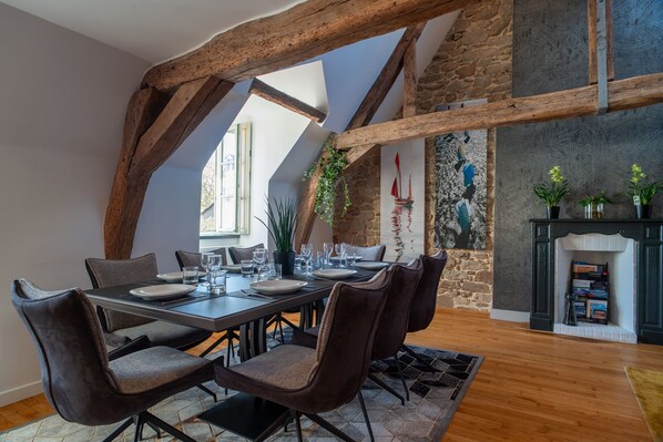 A dining area with a high ceiling, exposed wooden beams, a stone wall, and a fireplace.