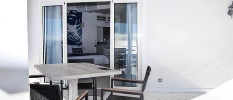 A balcony with a table and chairs, a sliding glass door leading to a bedroom, and a white exterior wall.