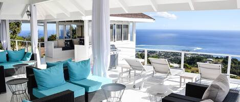 A covered outdoor seating area with wicker furniture and cushions, overlooking a scenic ocean view.