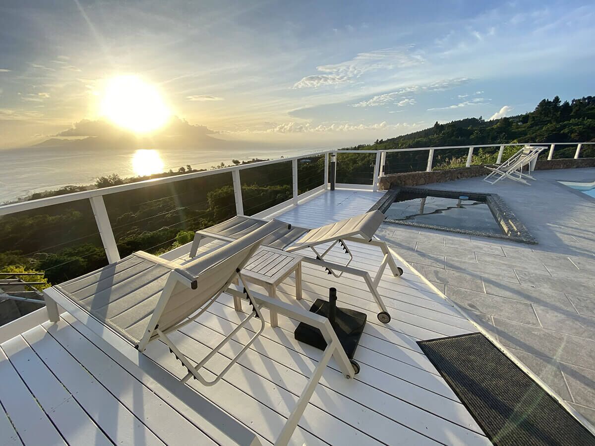 A sunlit deck with lounge chairs and a small table, overlooking a body of water and greenery.