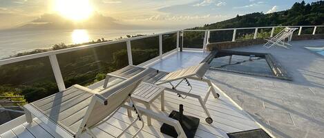 A sunlit deck with lounge chairs and a small table, overlooking a body of water and greenery.