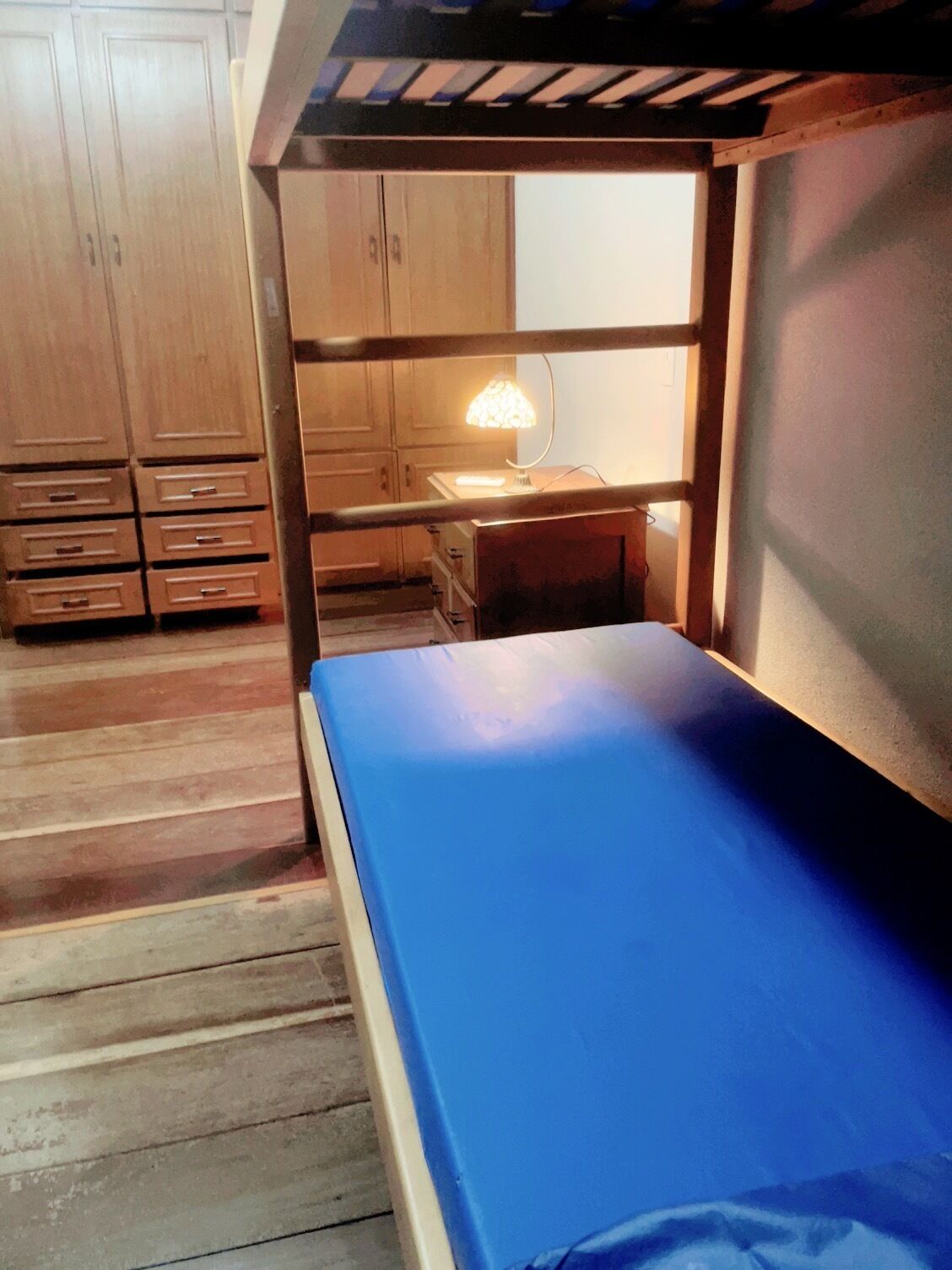 Una cama nido con colchón azul, armarios de madera y un escritorio con lámpara.