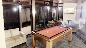 A pool table in a room with a tiled floor, a sofa, and a glass partition with a view of palm trees. - Beach House Unid. 3 Casas Praia Guarujá (Guarujá)
