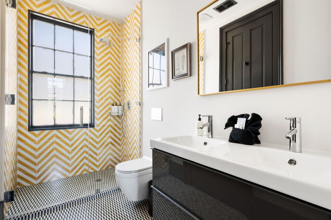 A bathroom with a chevron-patterned wall, a large window, a white sink with two faucets, and a black towel hanging on the sink.